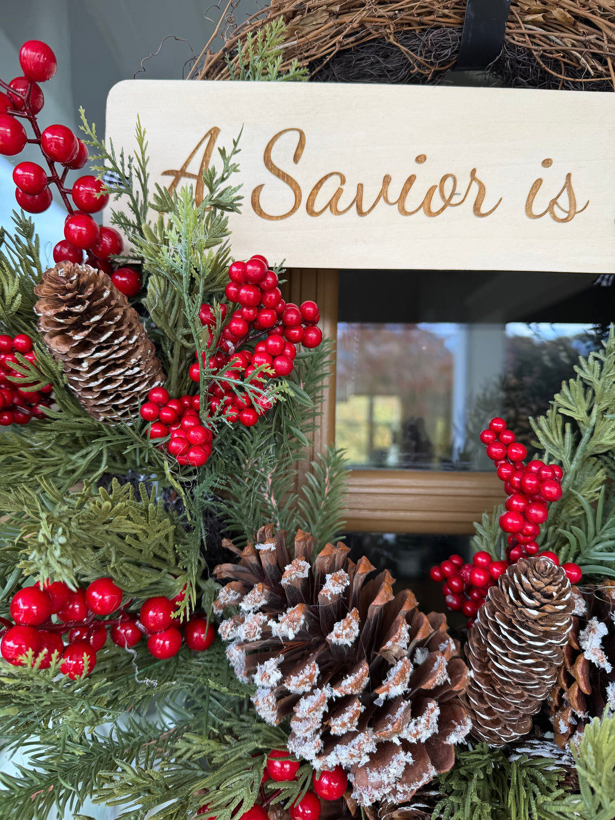 Sons & Daughters “A Savior Is Born” door wreath with festive Christmas greenery and decorative lettering